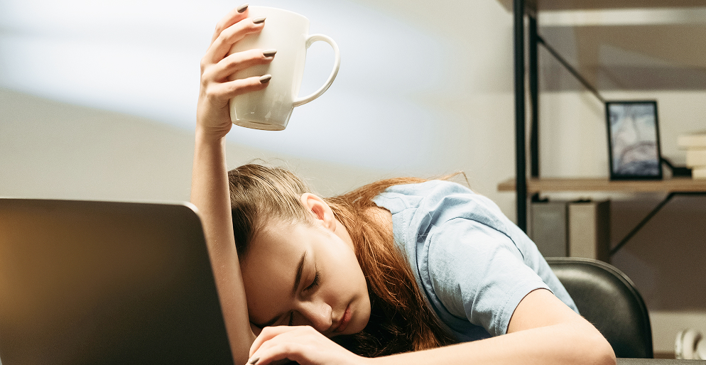Уставшая женщина с чашкой за рабочим столом — хроническая усталость и недосыпание как факторы нарушения микробиоты кишечника