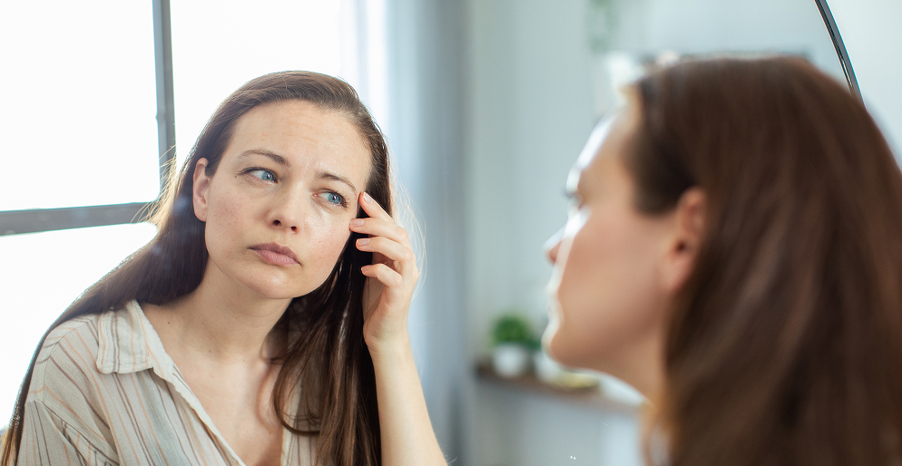 Проблемы с кожей при ПМС: связь дисбиоза кишечника и воспалений на лице Женщина рассматривает воспаления на коже перед зеркалом — влияние дисбиоза кишечника на состояние кожи при ПМС